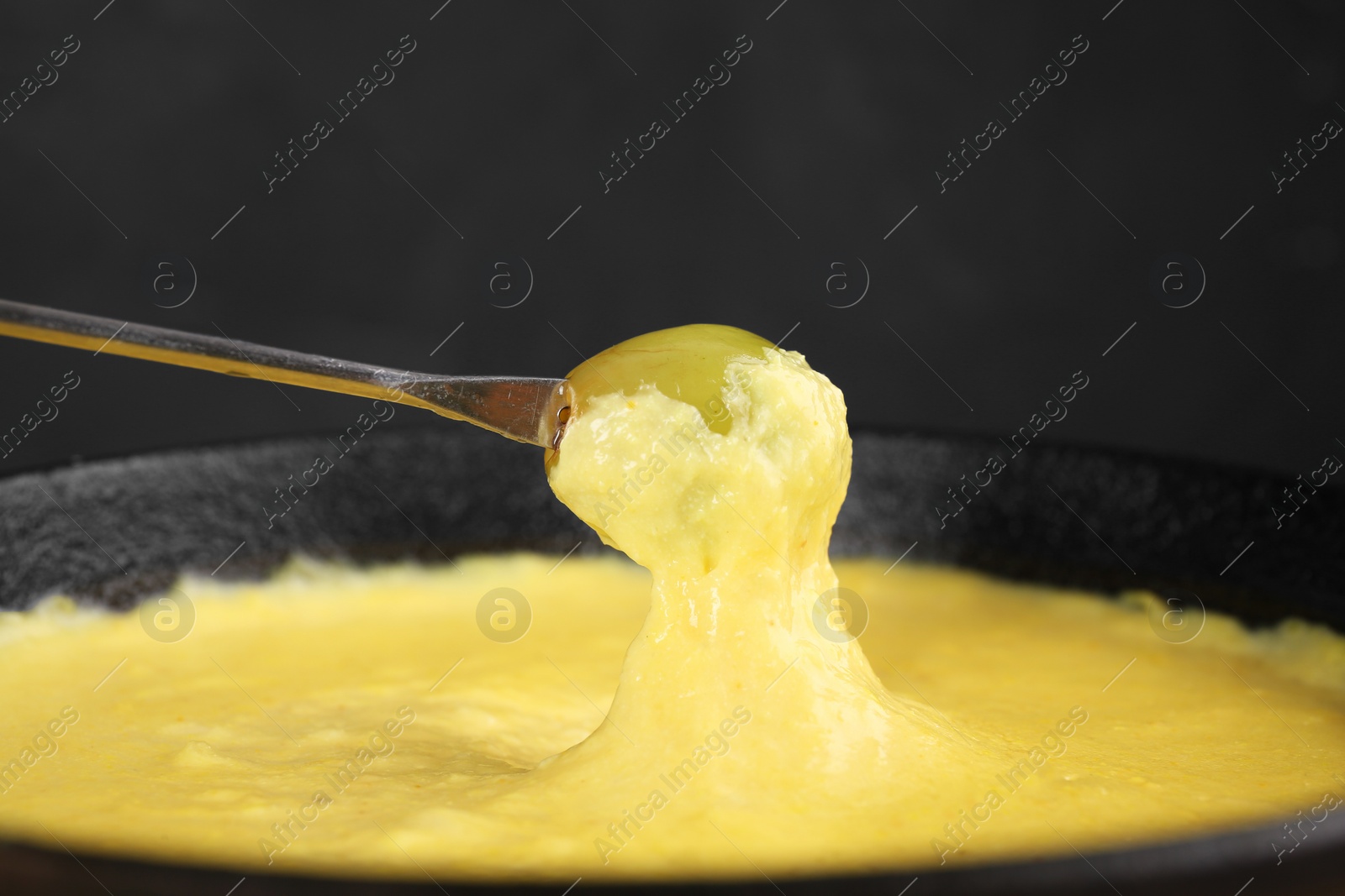 Photo of Dipping grape into fondue pot with tasty melted cheese against dark gray background, closeup