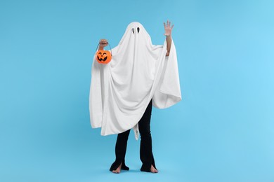Photo of Woman in white ghost costume holding pumpkin bucket on light blue background. Halloween celebration