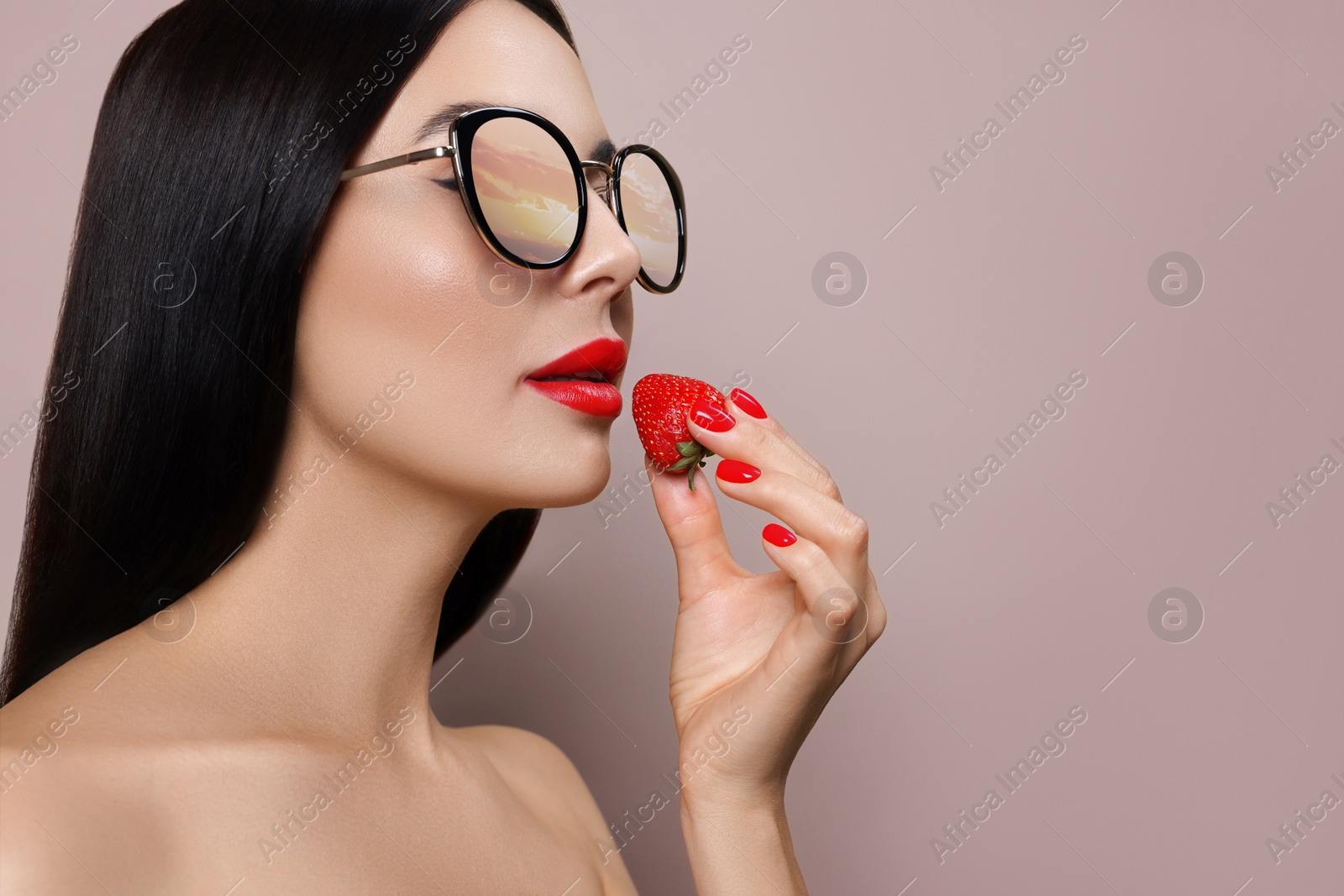 Image of Attractive woman in stylish sunglasses holding strawberry on grey beige background. Sunset sky reflecting in lenses. Space for text