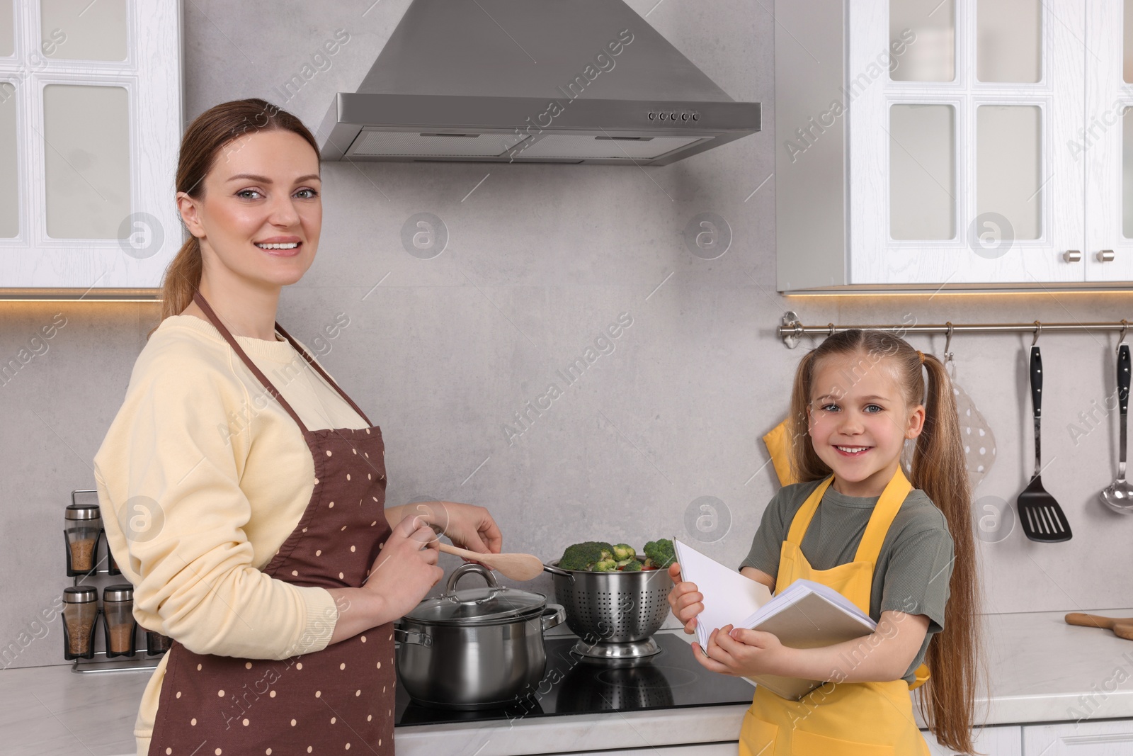 Photo of Cute little girl with her mother cooking by recipe book in kitchen