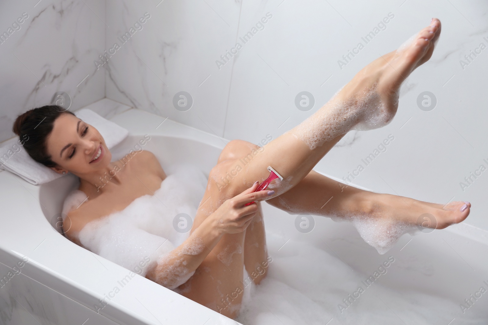 Photo of Young woman shaving legs while having bubble bath