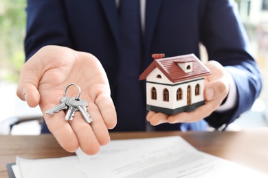 Photo of Real estate agent holding house model and keys at table in office, closeup