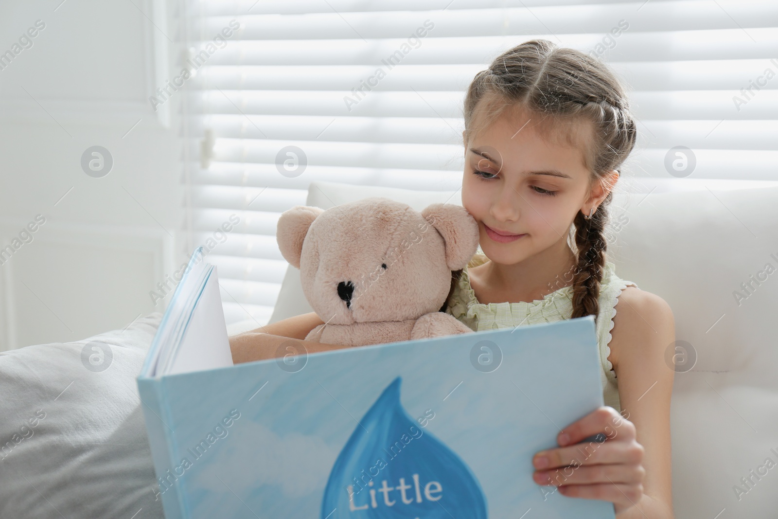 Photo of Cute little girl with teddy bear reading book on sofa at home