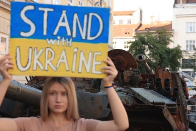 Photo of Sad woman holding poster in colors of national flag with words Stand with Ukraine near broken tank on city street