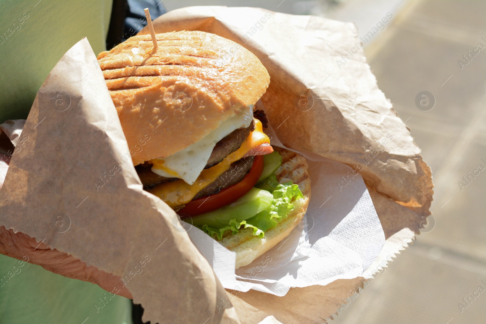 Photo of Woman holding delicious burger in paper wrap on blurred background, closeup