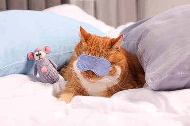 Photo of Cute ginger cat with sleep mask and crocheted mouse resting on bed