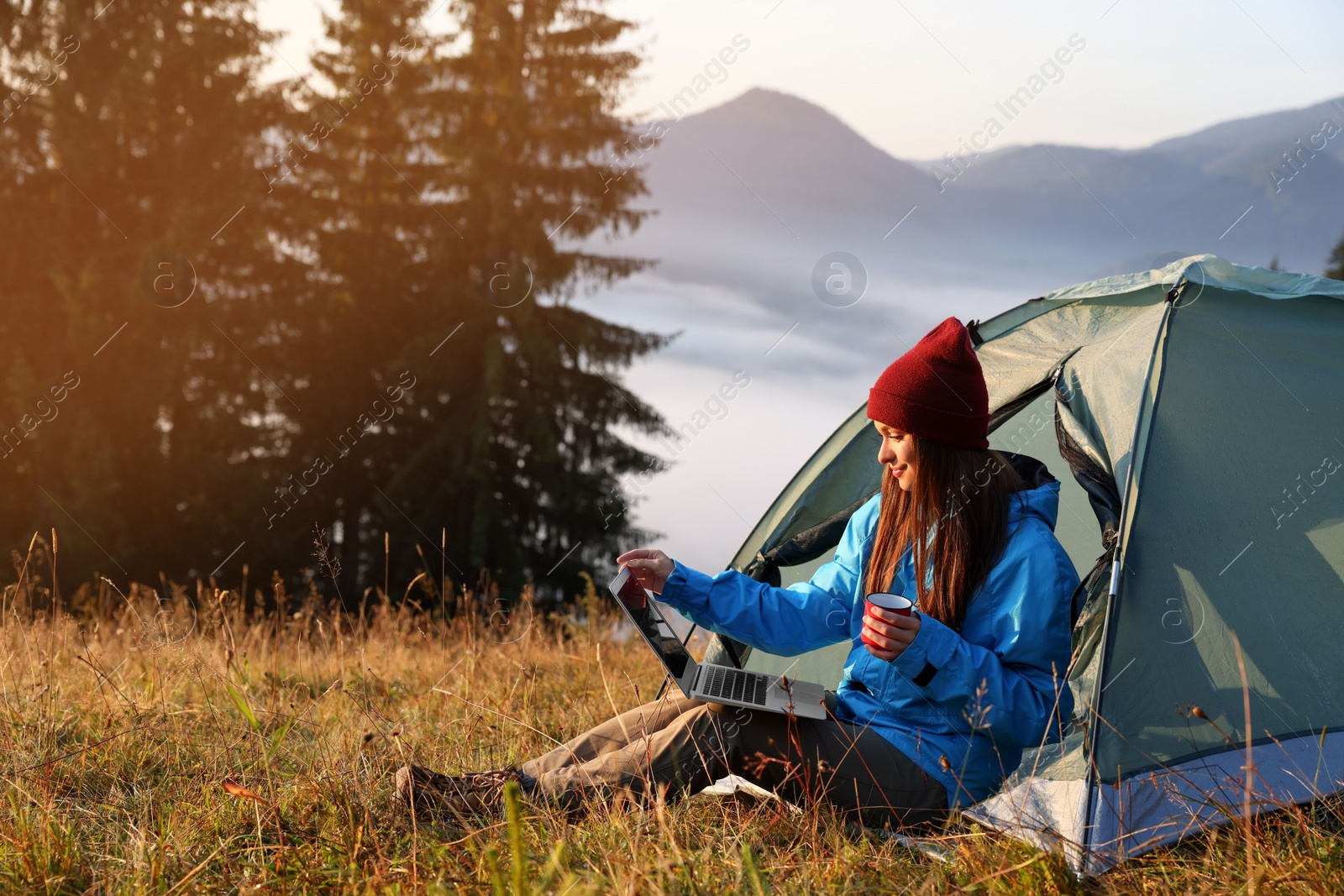 Photo of Woman working on laptop near camping tent outdoors surrounded by beautiful nature