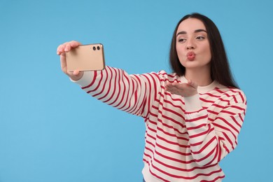 Young woman taking selfie with smartphone and blowing kiss on light blue background