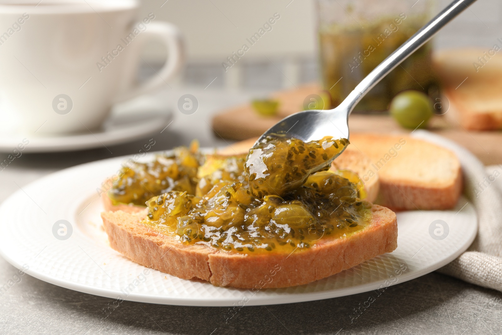 Photo of Spreading delicious gooseberry jam onto toast on grey table, closeup