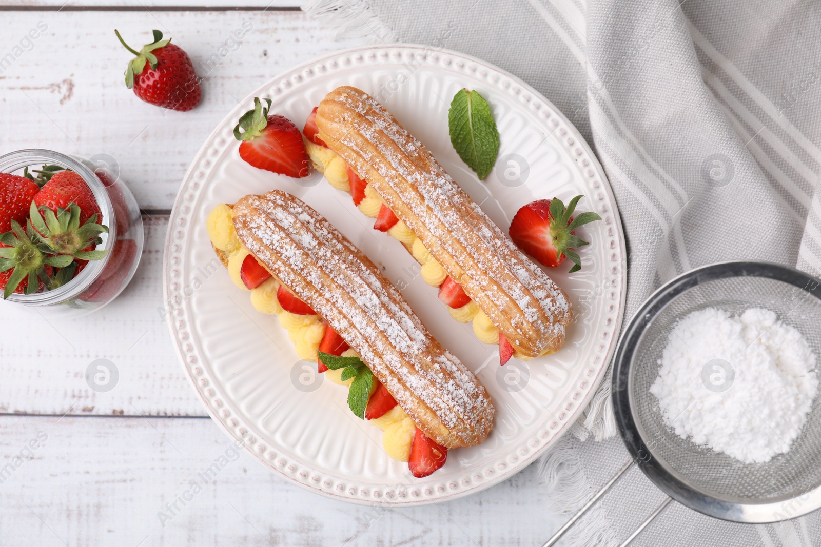 Photo of Delicious eclairs filled with cream, strawberries, mint and powdered sugar on white wooden table, top view