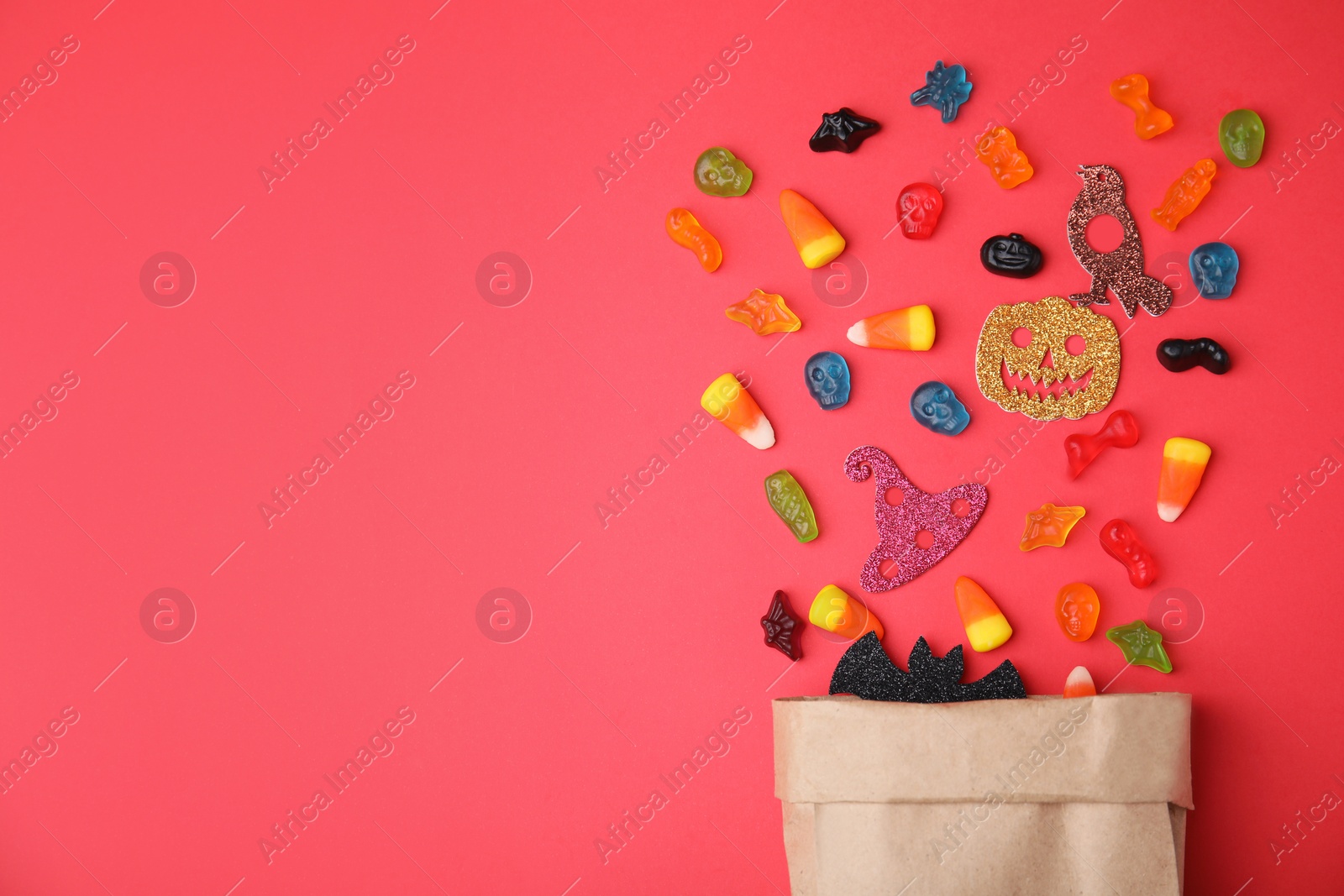 Photo of Paper bag of tasty candies and Halloween decorations on red background, flat lay. Space for text