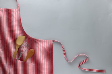 Photo of Red striped apron with kitchen tools on light grey background, top view. Space for text