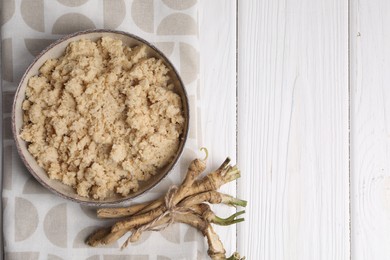 Bowl of tasty prepared horseradish and roots on white wooden table, flat lay. Space for text