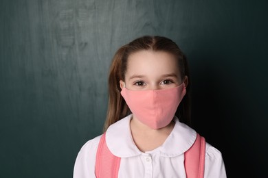 Photo of Little girl wearing protective mask and backpack near chalkboard. Child safety