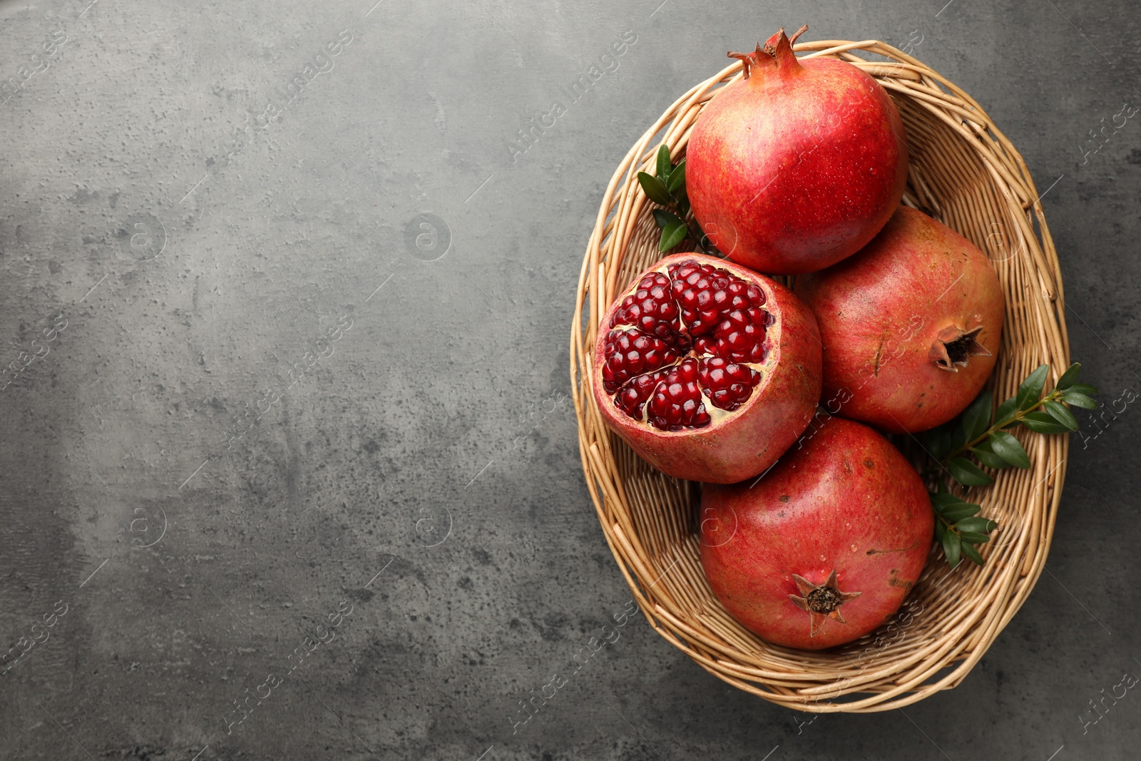 Photo of Fresh pomegranates and green leaves in wicker basket on grey table, top view. Space for text