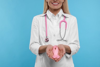 Doctor with stethoscope holding pink ribbon on light blue background, closeup. Breast cancer awareness