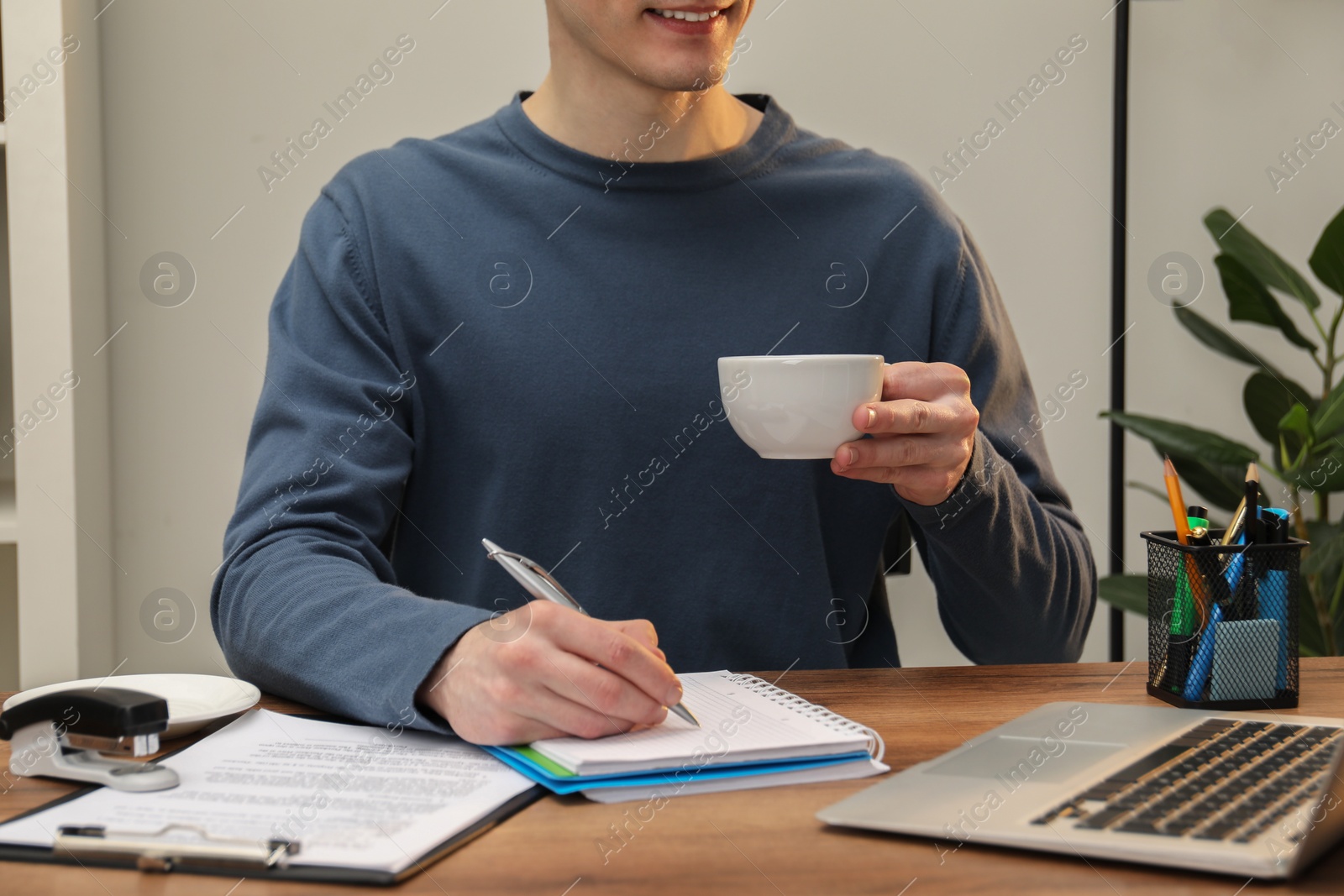 Photo of Man with cup of drink taking notes at wooden table in office, closeup