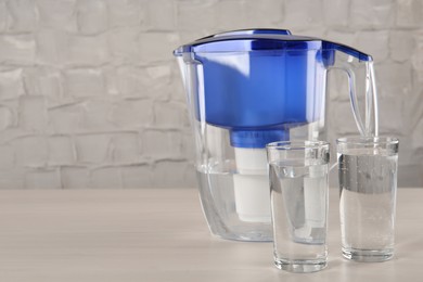 Photo of Filter jug and glasses with purified water on white table indoors. Space for text