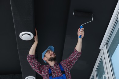 Photo of Worker in uniform painting ceiling with roller indoors, low angle view