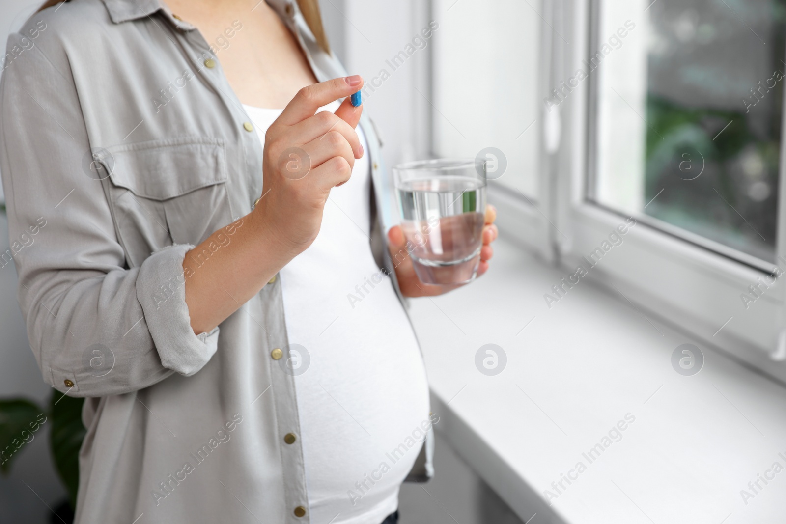 Photo of Pregnant woman holding pill and glass of water near window indoors, closeup