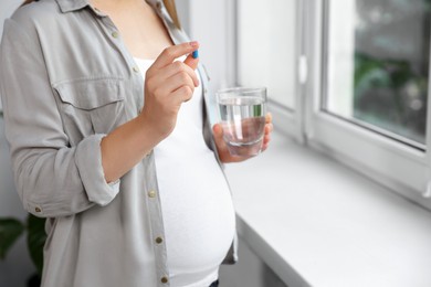 Photo of Pregnant woman holding pill and glass of water near window indoors, closeup