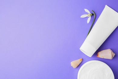 Jar and tube of cream, seashells with snowdrop flower on violet background, flat lay. Space for text