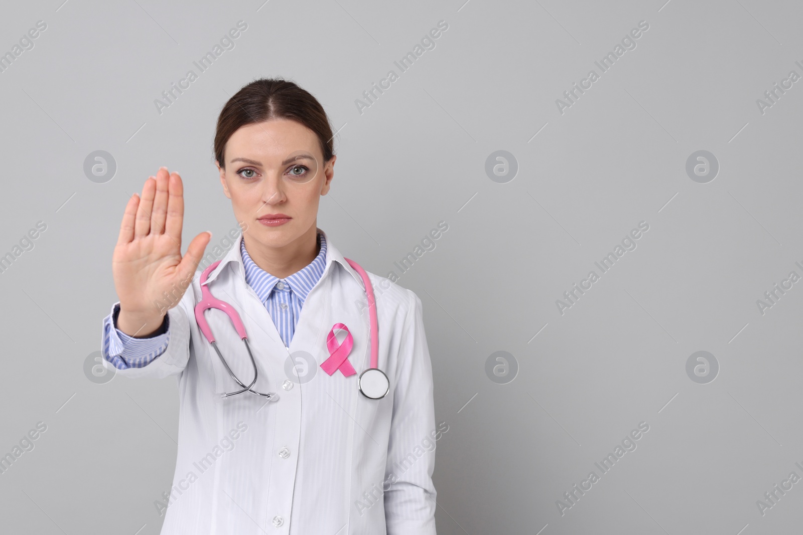 Photo of Mammologist with pink ribbon showing stop gesture against grey background, space for text. Breast cancer awareness