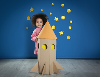 Cute African American child playing with cardboard rocket near blue wall