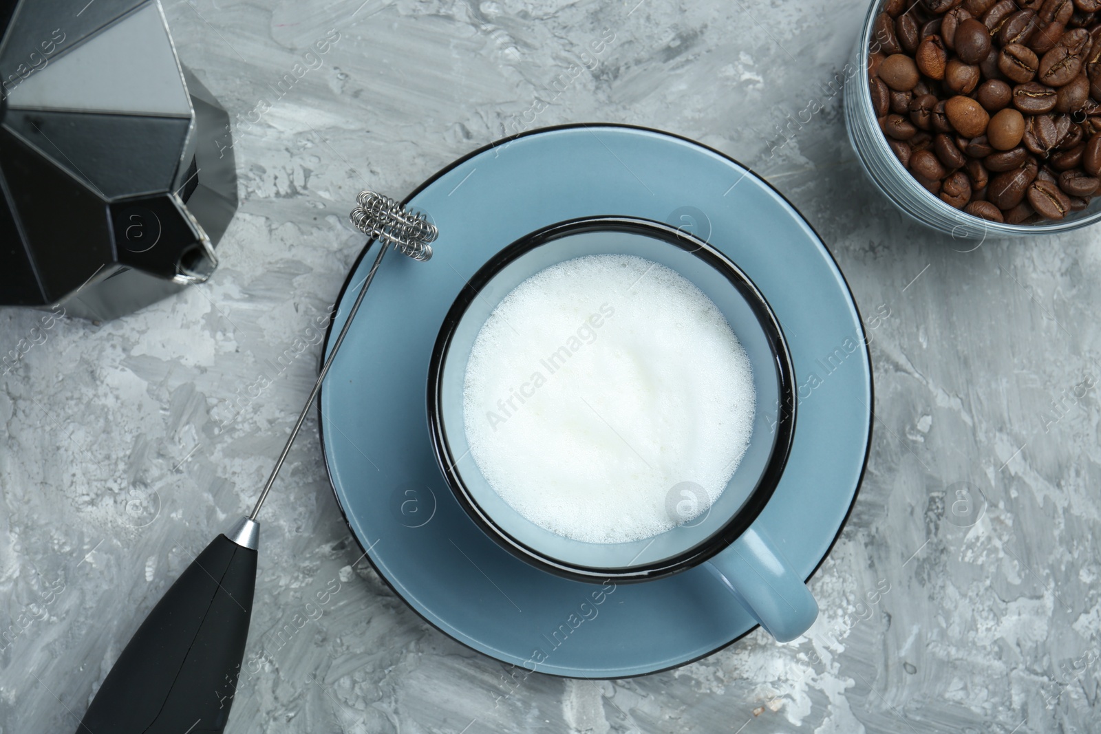 Photo of Mini mixer (milk frother), whipped milk in cup, coffee beans and moka pot on grey textured table, flat lay