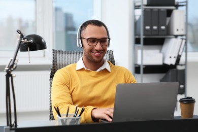 Photo of Young man with headphones working on laptop at table in office