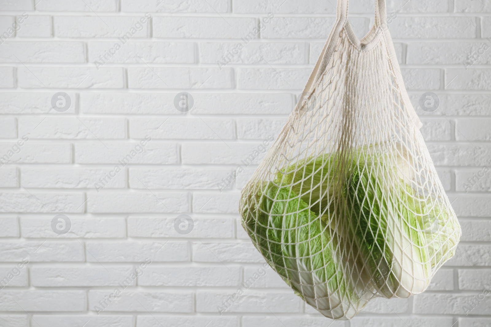 Photo of Woman holding string bag with fresh Chinese cabbages near white brick wall, closeup. Space for text