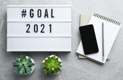 Photo of Light box with hashtag Goal 2021 near notebooks and modern smartphone on grey table, flat lay. New year targets