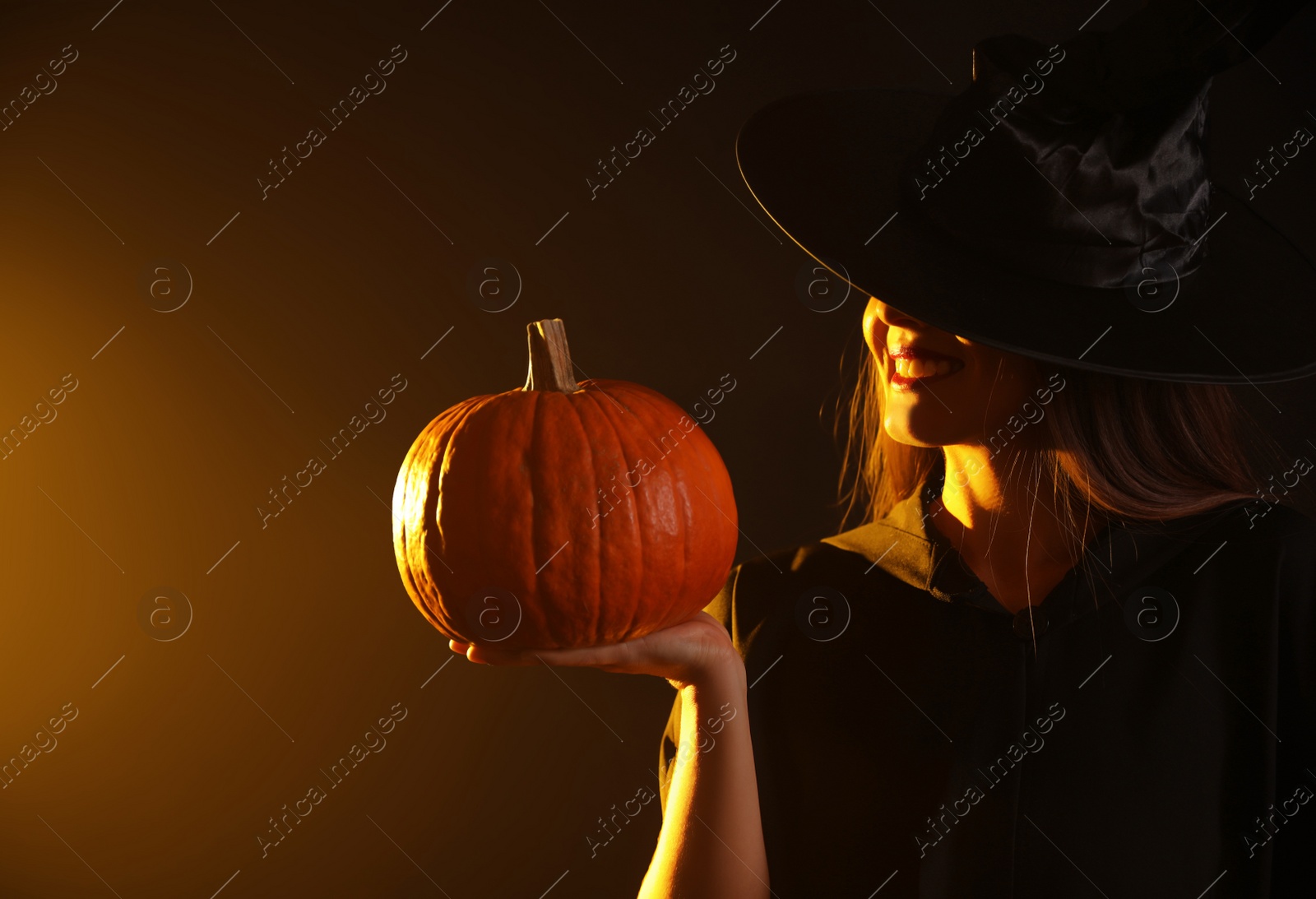 Photo of Young woman wearing witch costume with pumpkin on dark background. Halloween party