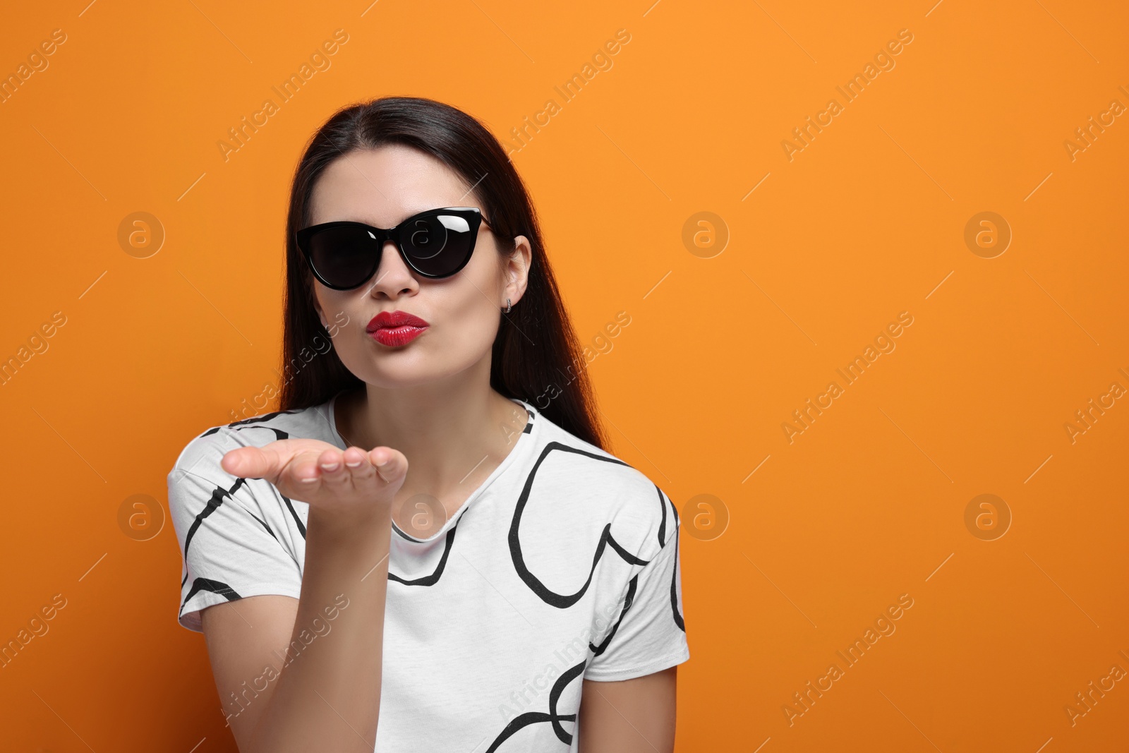 Photo of Beautiful young woman in stylish sunglasses blowing kiss on orange background. Space for text