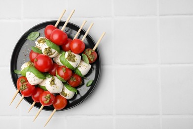 Caprese skewers with tomatoes, mozzarella balls, basil and pesto sauce on white tiled table, top view. Space for text