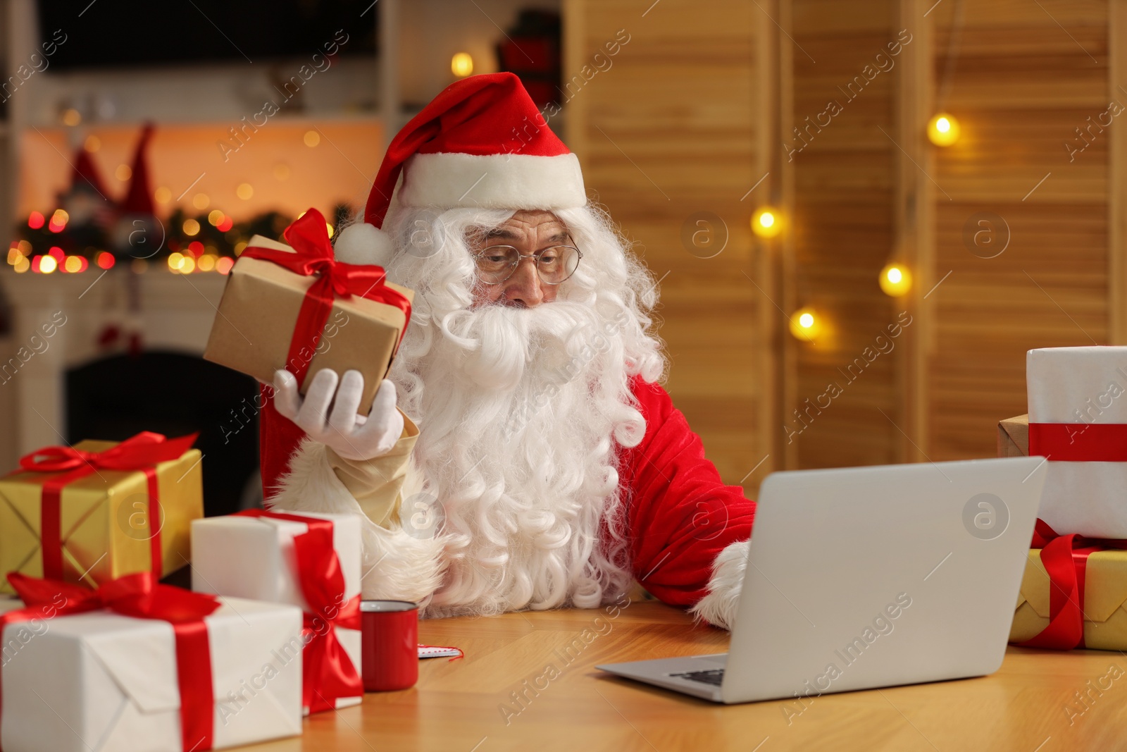 Photo of Santa Claus with Christmas gift using laptop at table in room