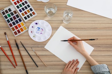 Photo of Woman painting with watercolor on blank paper at wooden table, top view