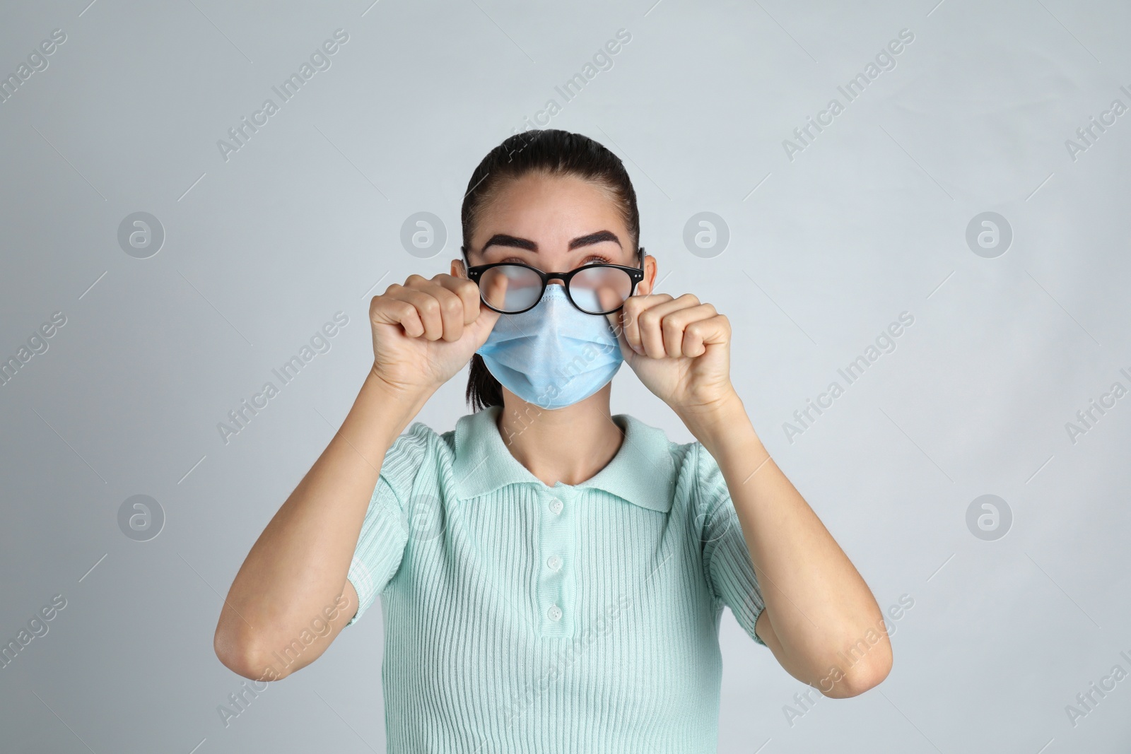 Photo of Woman wiping foggy glasses caused by wearing medical mask on light background