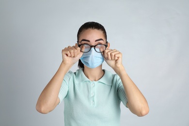 Photo of Woman wiping foggy glasses caused by wearing medical mask on light background