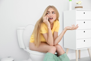 Young woman with mobile phone sitting on toilet bowl at home