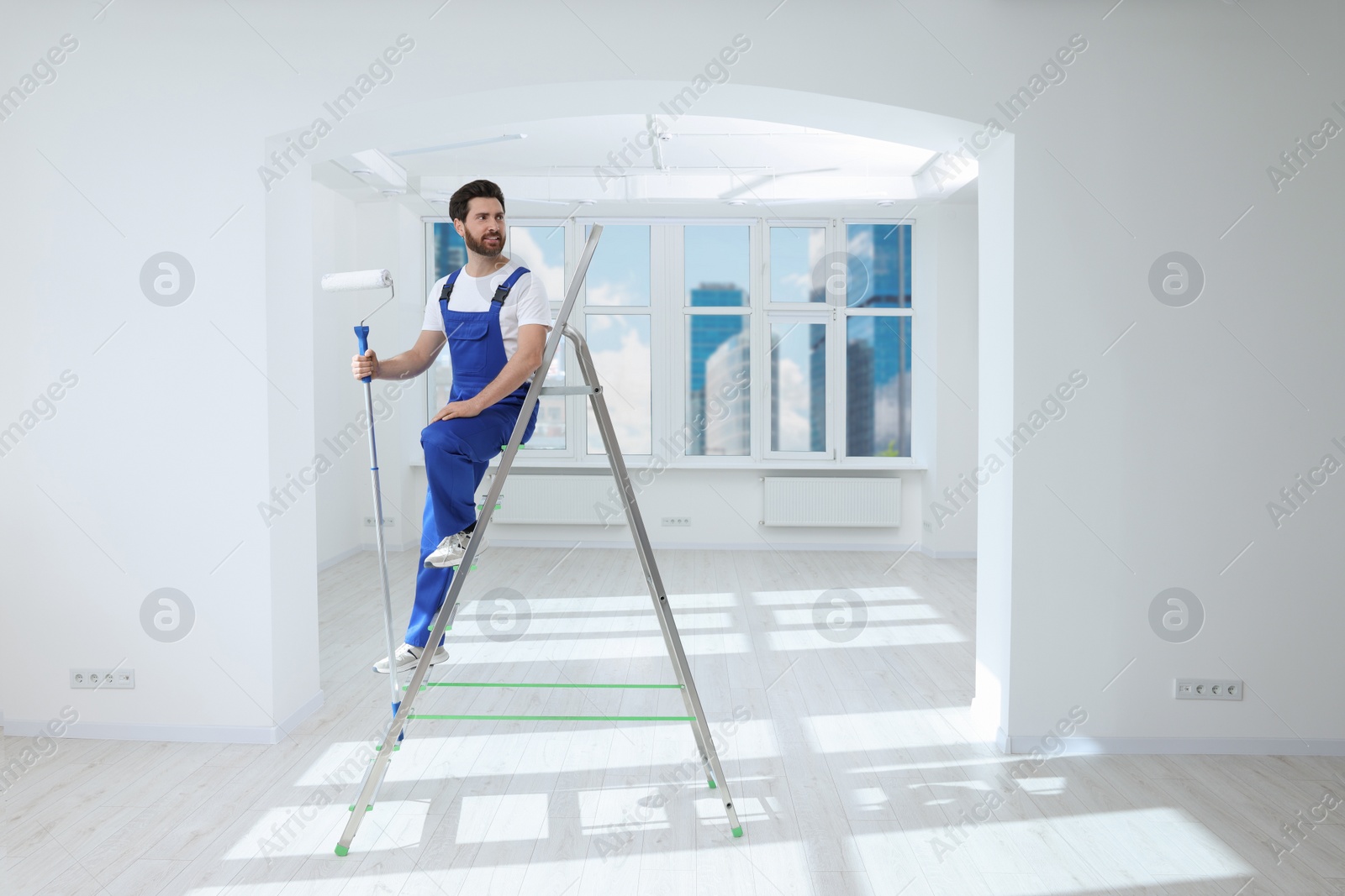 Photo of Handyman with roller on step ladder in room. Ceiling painting