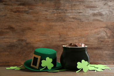 Pot of gold coins, hat and clover leaves on wooden table, space for text. St. Patrick's Day celebration