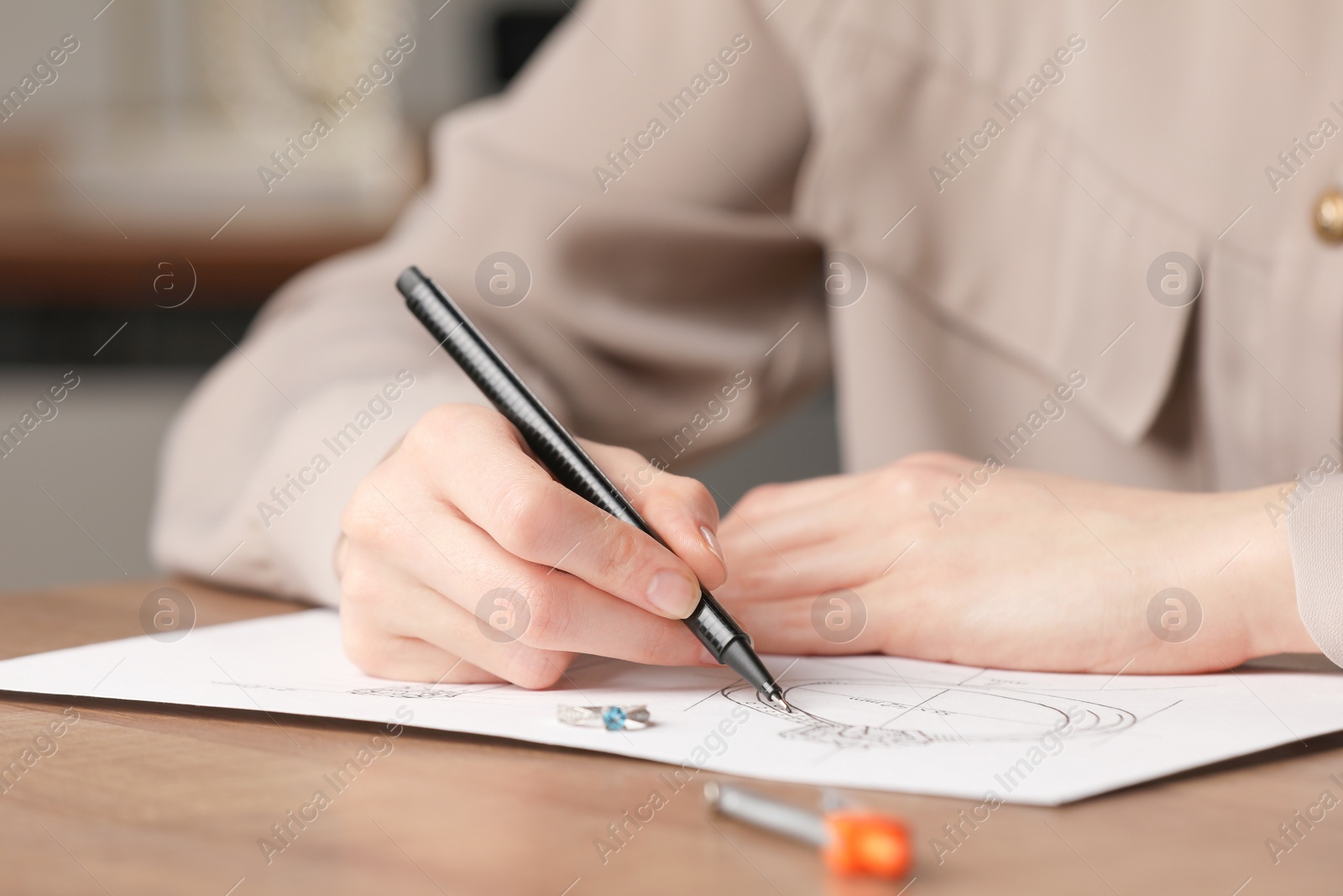 Photo of Jeweler drawing sketch of elegant ring on paper at wooden table indoors, closeup