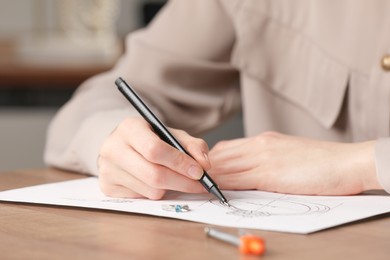 Jeweler drawing sketch of elegant ring on paper at wooden table indoors, closeup