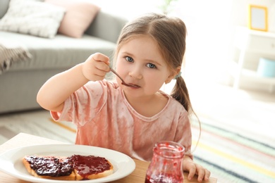 Photo of Little girl eating jam at table in living room