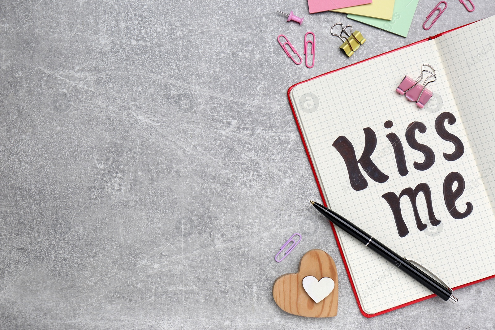 Photo of Phrase Kiss me written in notebook and stationery on light grey stone table, flat lay. Space for text