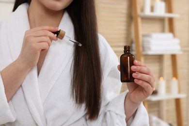 Photo of Woman with bottle applying essential oil onto hair in bathroom, closeup