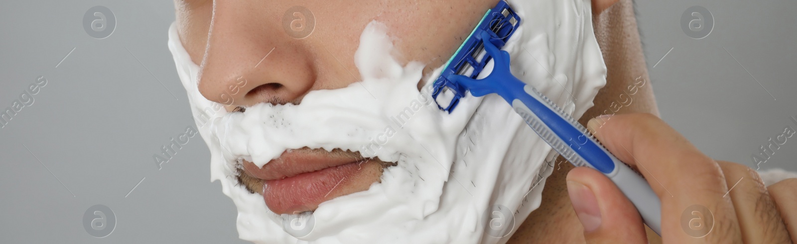 Image of Young man shaving with razor on grey background, closeup. Banner design