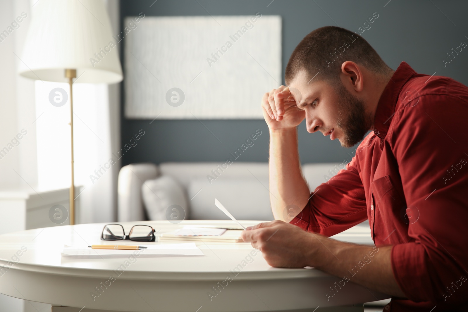 Photo of Worried man reading paper letter at white table in room