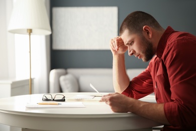 Worried man reading paper letter at white table in room